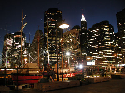New York skyline from the piers of the Fulton Market area. Tall ships sway in the harbor, while a couple embraces on the bench